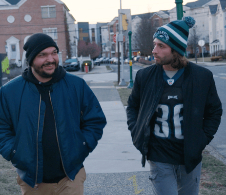 TRNN Editor-in-Chief Maximillian Alvarez smiles while walking with Home Depot Employee Vince Quiles in Philadelphia, PA. They are wearing jackets and walking along a sidewalk. Residential buildings and a grey sky can be seen in the background.